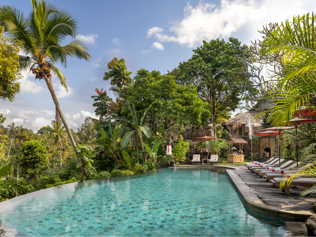 An inviting photograph capturing the main infinity pool area at Puri Gangga Resort, set against a lush, dense tropical jungle background under a partly cloudy blue sky. The large pool features clear turquoise water with a stone deck to the right. On the deck, there is a row of wooden sun loungers with crisp white towels and vibrant red decorative pillows, positioned under matching red umbrellas. In the distance, traditional thatched-roof villas and an open-air bar/dining area can be seen among the palm trees.